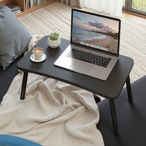 Könnte beinhalten: Ein schwarzer Laptop-Tisch mit einem Laptop, einer Kaffeetasse und einer kleinen Sukkulentenpflanze. Der Laptop zeigt eine Strandszene. Der Tisch hat schwarze Beine und steht auf einem Sofa mit einer Decke. Tischmaße in cm.