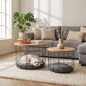 Könnte beinhalten: Zwei runde Couchtische mit Holzplatten und schwarzen Drahtkäfig-Untergestellen. Auf einem Tisch liegt ein kleiner weißer Hund auf einem grauen Kissen. Der andere Tisch hat ein graues Kissen. Die Tische stehen in einem Wohnzimmer.