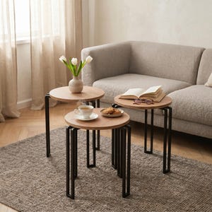 Könnte beinhalten: Ein Satz von drei runden Beistelltischen mit hellen Holzplatten und schwarzen Metallbeinen. Auf einem Tisch steht eine Vase mit weißen Tulpen, auf einem anderen eine Teetasse und Kekse, und auf dem dritten ein offenes Buch und eine Brille.