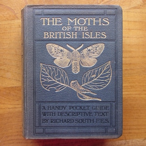 Könnte beinhalten: Ein Vintage-Buch mit dem Titel "The Moths of the British Isles" mit goldener Schrift und Illustrationen einer Motte und Blättern auf einem dunkelblauen Einband. Ein praktischer Taschenführer mit beschreibendem Text.