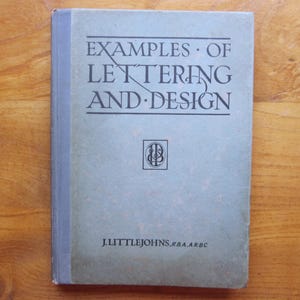 Op de afbeelding: Een gebonden boek met de titel Examples of Lettering and Design door J. Littlejohns, R.B.A., A.R.B.C. De kaft is gedempt lichtblauw-grijs met donkerblauwe tekst en een centraal vierkant embleem met de in elkaar verweven letters J en L.