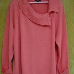 May include: A coral-colored sweater with a unique asymmetrical collar design. The long-sleeved top features ribbed cuffs and a smooth, solid fabric. The sweater is hanging on a white hanger against a light brown background.