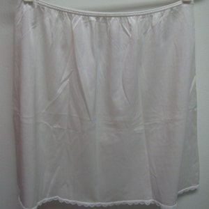 May include: A white half slip hanging on a white hanger. The slip is made of a smooth, silky fabric and has a scalloped lace trim at the hem. The elastic waistband is gathered.