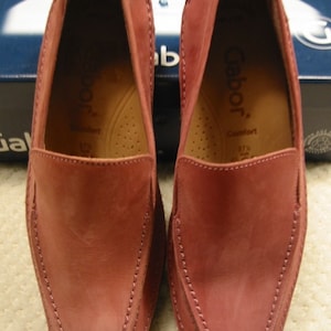 May include: A pair of dusty rose-colored Gabor loafers. The shoes have a square toe design with visible stitching along the seams. The interior of the shoe is a lighter shade of brown, with the brand name "Gabor" visible. The shoes are size 37 1/2.