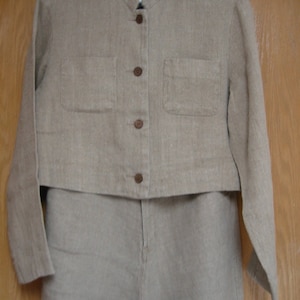 May include: A two-piece beige linen suit. The jacket has a mandarin collar, two front pockets, and brown buttons. The pants are high-waisted and straight-legged. The suit is hanging on a white hanger against a wooden door.