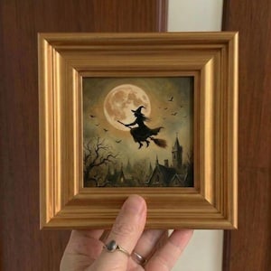 May include: A square painting in a gold-toned frame depicting a silhouette of a witch flying on a broomstick against a large, pale yellow full moon. Below the moon, dark silhouettes of bare trees and a gothic-style building are visible.