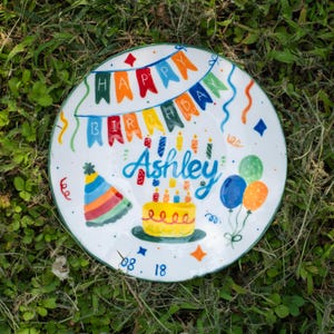 May include: A white ceramic plate featuring a hand-painted birthday theme with a colourful bunting banner reading "HAPPY BIRTHDAY", the name "Ashley" in blue script, a yellow cake with candles, a striped party hat, balloons, and "08.18".