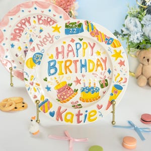 Puede incluir: Un plato de cerámica blanca con decoraciones en relieve de colores, que incluye el texto "HAPPY BIRTHDAY" en azul y amarillo, el nombre "Katie" en naranja y un icono de calendario que muestra "May 22". El diseño incluye pasteles y estrellas.
