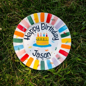 May include: A white ceramic plate featuring a hand-painted birthday cake illustration in the centre with the text Happy Birthday Jason and the date 08 18 2023. The rim is decorated with alternating colourful rectangular brush strokes.