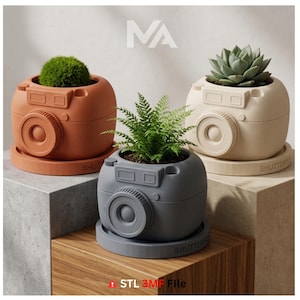 May include: Three camera-shaped planters in terracotta, cream, and gray, each holding a different plant. The planters have a lens and shutter button design, with the word "SHUTTER" on the base. The plants include a succulent, fern, and moss.