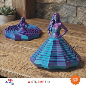 Könnte beinhalten: Zwei 3D-gedruckte Fidget-Spielzeuge in Form einer weiblichen Figur mit langem Haar und Händen in den Hüften. Die Figuren haben ein violett-türkises Farbverlauf-Finish. Eine Figur steht auf einem gestuften, breiten, geometrischen Rock, die andere sitzt in einer flachen Basis mit Spiralmuster. Text: 3D Fidget, STL Craft Hub, STL 3MF File.