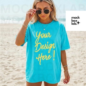 Puede incluir: Una camiseta turquesa con el texto "Your Design Here!" en escritura amarilla. La camiseta la lleva una persona en una playa, con gafas de sol y joyas. El logo de mock box lab está en la esquina superior derecha.
