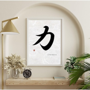 May include: A framed print featuring a black Japanese calligraphy symbol on a textured white background. The word "STRENGTH" is printed below the symbol. The print is displayed on a wooden shelf with a gold lamp, clock, and vase of dried flowers.