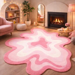 Könnte beinhalten: Ein rosa-weißer Teppich mit einer einzigartigen, fließenden Form. Der Teppich hat ein Farbverlaufsdesign, das von einem tiefrosa Rand zu helleren Rosa- und Weißtönen in der Mitte übergeht. Der Teppich liegt auf einem Holzboden.