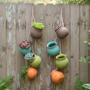 May include: A collection of hanging ceramic planters in various colors, including brown, teal, green, and orange, suspended by rope against a weathered wooden fence. Each planter holds different types of small plants and succulents, creating a vertical garden display.