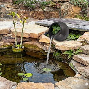 Puede incluir: Una fuente de agua de cerámica negra con agua que fluye hacia un pequeño estanque. La fuente está rodeada de rocas, plantas y un puente de madera. El agua fluye desde una fuente redonda en forma de cuenco hacia el estanque, creando ondulaciones.