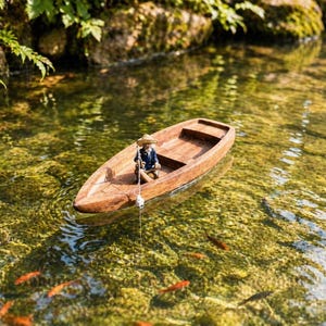 Pode incluir: Um pequeno barco de madeira com uma figura em miniatura de um pescador com chapéu de palha e camisa azul flutua na água. O barco é cercado por peixes em um lago com algas verdes e rochas.