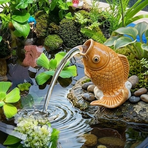 Könnte beinhalten: Ein Keramik-Fischbrunnen, aus dessen Maul Wasser in einen kleinen Teich fließt. Der Fisch hat eine warme orange Farbe mit detaillierten Schuppen. Der Teich ist von üppigen grünen Pflanzen und kleinen Steinen umgeben.