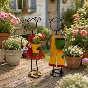 Könnte beinhalten: Zwei Metall-Gartenfiguren in Strichmännchen-Optik. Eine Figur trägt ein rotes Kleid mit weißen Blumen-Ausschnitten und hält einen gelben Eimer. Die andere trägt eine gelbe Latzhose und hält einen grünen Eimer. Beide haben schwarze Drahtrahmen.