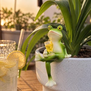 Puede incluir: Figura de rana verde con ojos amarillos, sosteniendo un vaso de líquido amarillo, posada en una maceta blanca. Adorno de jardín divertido y peculiar, ideal para un ambiente relajado.