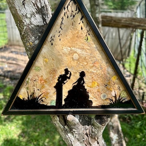May include: A triangular framed artwork featuring a silhouette of a couple in formal attire. The background has a textured, light-colored surface with floral and leaf accents. The frame is black, and the artwork is displayed outdoors.