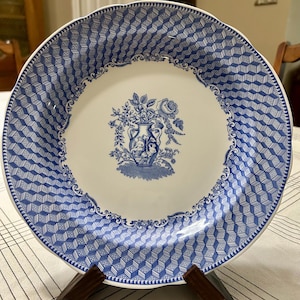 May include: A white and blue decorative plate with a floral design in the center. The plate has a geometric pattern around the rim and is displayed on a wooden stand. The plate is approximately 10 inches in diameter.