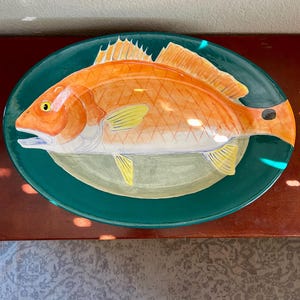 May include: An oval ceramic serving platter featuring a hand-painted orange fish with yellow fins and a black spot near the tail. The fish is centered on a deep teal background, with a light green oval area directly beneath the fish body.