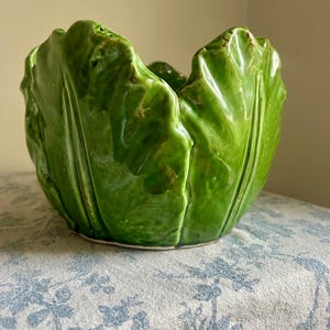 May include: A glossy, bright green ceramic bowl shaped like a head of lettuce. The exterior features realistic, overlapping leaf textures with raised veins and ruffled edges, creating a deep, rounded vessel with a smooth, shiny finish throughout.