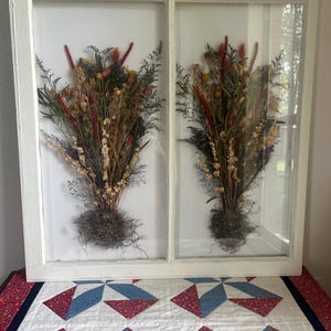 Puede incluir: Una exhibición en una ventana con marco blanco que presenta dos arreglos de flores secas. Los ramos incluyen elementos rojos, amarillos y marrones, con follaje verde y una base de musgo gris. La ventana se encuentra sobre un edredón con patrones geométricos rojos, blancos y azules.