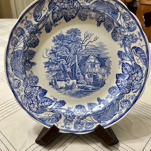 May include: A blue and white decorative plate with a scalloped edge. The plate features a detailed landscape scene in the center and a floral border. The plate is displayed on a wooden stand.
