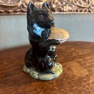 May include: A glossy black ceramic figurine of a dog holding a plate with a light-colored pastry. The dog wears a blue bow tie. The base is green and the figurine is on a wooden surface.