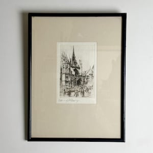 May include: A framed monochrome etching depicting a cathedral with a prominent spire and detailed architecture. The artwork is presented on a cream mount, encased in a black frame. The image includes a signature at the bottom.