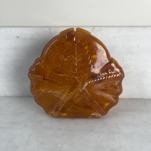 May include: An antique amber-toned clutch bag with a leaf motif. The bag has a rounded form and a textured finish. The words "Mich Repa" are written on the side in cursive. The clasp is at the top.