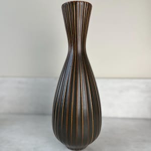 May include: A tall, dark brown vase with vertical ribbed detailing. The vase has a slender neck that widens to a bulbous body. The ribbed design is accented with a lighter brown colour, creating a contrast. The vase is likely ceramic or stoneware.