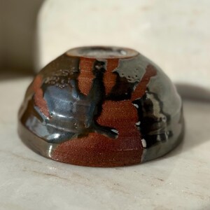 Peut inclure: Un petit bol en céramique fait à la main avec un glaçage unique. Le bol présente un mélange de couleurs, notamment le brun rougeâtre, le noir et le gris, créant un aspect texturé et organique. La forme du bol est arrondie, avec un bord légèrement évasé.