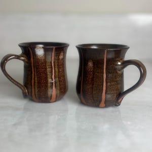 May include: Two handmade ceramic mugs with a dark brown glaze and vertical copper-coloured stripes. Each mug has a curved handle and a slightly flared rim. The mugs are displayed on a white surface.