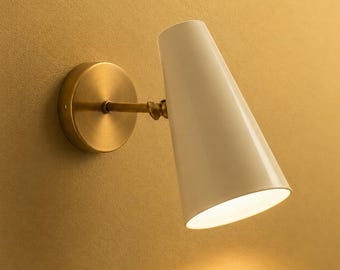 May include: A wall-mounted lamp featuring a conical white shade attached to a circular brass-toned base by a short horizontal arm. The fixture is angled downwards, casting a soft light from the open bottom of the white metal cone structure.