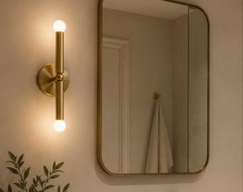 May include: A bathroom scene featuring a gold-toned wall sconce with two spherical lights, a rectangular mirror with rounded corners, and a gold faucet. A vase with greenery and a bottle of soap are visible.