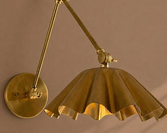 May include: A wall-mounted lamp with a brass finish, featuring a circular wall plate, an adjustable articulated arm with two pivot points, and a wide, scalloped, bell-shaped shade that directs light downwards from its metallic interior surface.