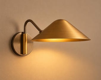 May include: A brass wall sconce with a conical shade. The sconce features a circular base and a curved arm supporting the shade. The shade is angled downwards, directing light. The fixture has a warm, golden colour, suitable for interior lighting.