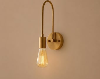 May include: A brass wall light with a curved arm and a circular base. It features a teardrop-shaped light bulb, emitting a warm, yellow light. The fixture is mounted on a neutral wall, creating a minimalist design. The overall design is simple and elegant.
