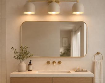 May include: A modern bathroom vanity with a rectangular mirror and a three-light fixture. The vanity features a light wood cabinet, a white countertop, and gold-toned hardware. A vase with greenery and a towel ring complete the design.