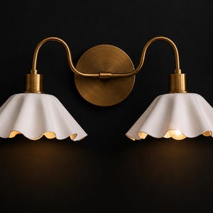 May include: A double-light wall sconce with a brass finish. The fixture features two white, scalloped-edge shades and a circular backplate. The sconce is mounted on a dark wall, with soft lighting emanating from the shades.