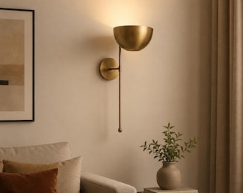 May include: A brass wall sconce with a half-bowl shade, a long stem, and a round wall mount. The sconce is mounted on a beige wall. A small round table with a vase of greenery and books is in the foreground.