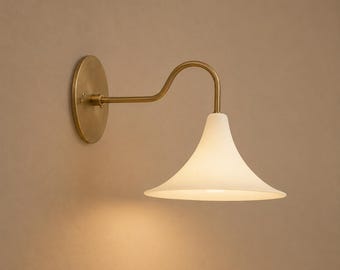 May include: A wall-mounted light fitting featuring a circular brass backplate and a curved brass arm that supports a wide, flared, matte white glass shade. The lamp emits a soft, warm glow from beneath the shade, highlighting the metal finish.