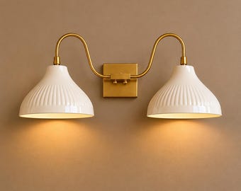 May include: A double wall light fixture featuring a gold-coloured metal frame and two white, ribbed lampshades. The sconce is mounted on a square gold-coloured base. The lampshades are lit, emitting a warm light.