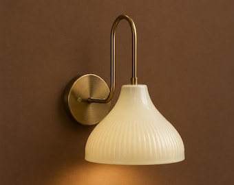 May include: A wall-mounted light fitting featuring a cream-coloured, ribbed lampshade and a brass-toned metal frame. The fitting has a circular base and a curved arm. The light is on, emitting a warm glow.