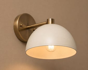 May include: A wall-mounted light fitting featuring a cream-coloured, dome-shaped lampshade. It has a brass-coloured base and arm, and a visible lightbulb. The design is minimalist and modern, suitable for interior lighting.