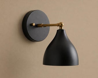 May include: A wall sconce featuring a matte black circular wall plate and a matching bell-shaped shade. The two components are connected by a horizontal brass-toned metal arm with a small cylindrical knob on the side of the wall plate.