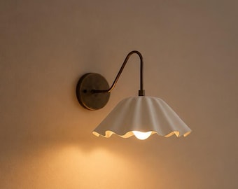 May include: A wall-mounted light fitting featuring a curved, dark bronze-toned metal arm attached to a circular base. The lamp includes a wide, off-white, scalloped-edge shade that directs light downwards, illuminating a single warm-toned light bulb.